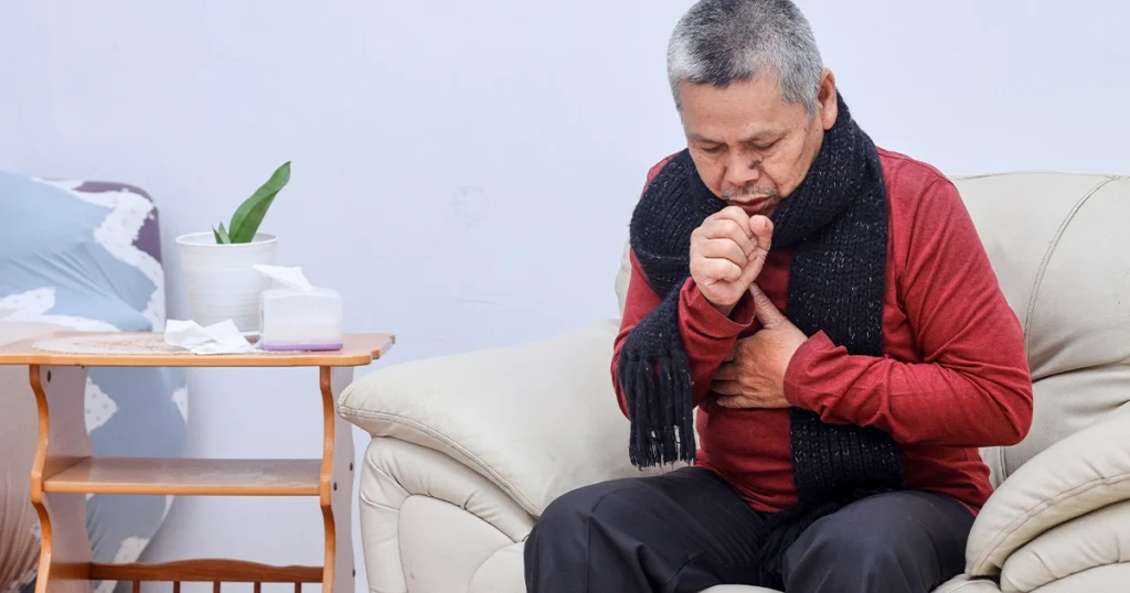 Elderly man coughing indoors showing common pneumonia symptoms.