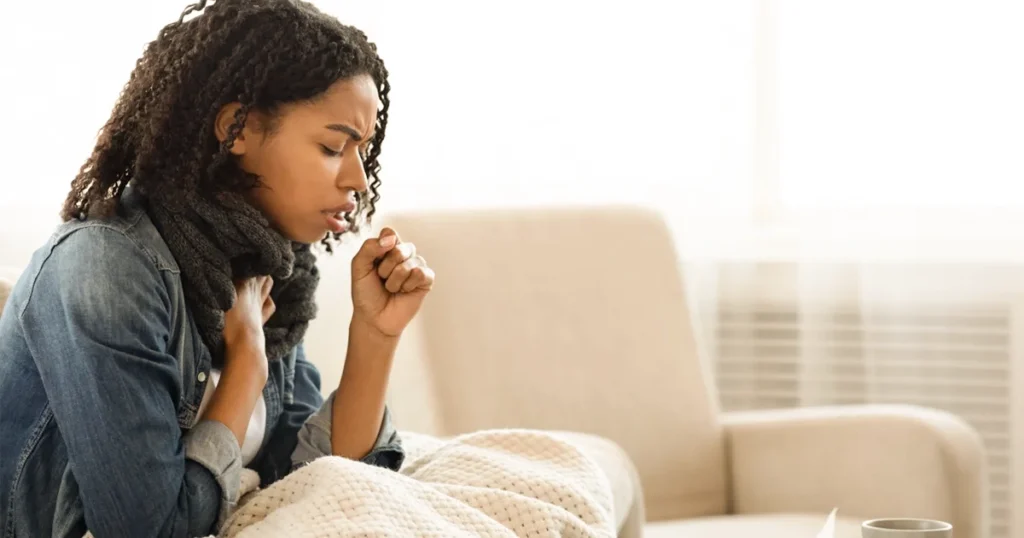 Young woman coughing indoors while showing mild pneumonia symptoms.