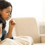 Young woman coughing indoors while showing mild pneumonia symptoms.