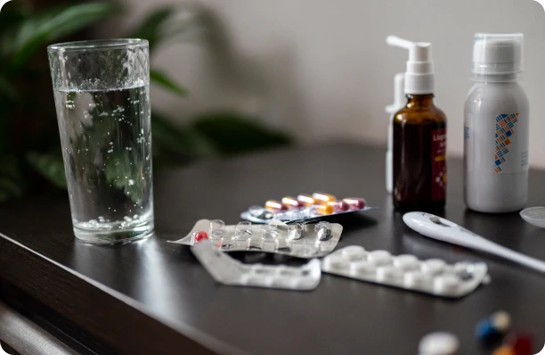Glass of water, medicine blister packs, bottles, and a thermometer on a table.