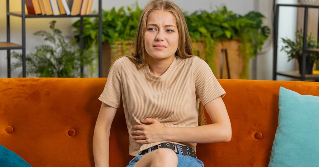 Young woman sitting on couch with visible bloating discomfort.